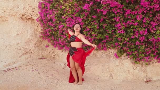 En brunett kvinna i en röd orientalisk dansdräkt med en sjal dansar mot bakgrund av blommande bougainvilleas — Stockvideo