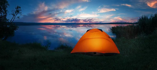 Barraca laranja iluminada internamente na costa do lago sob o dramático céu do pôr-do-sol — Fotografia de Stock