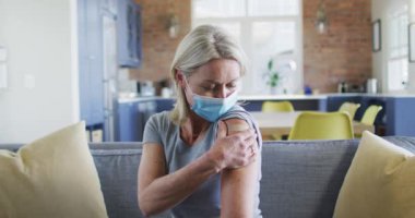 Oturma odasında kolunda bandajla yüz maskesi takan mutlu beyaz kadın portresi. covid 19 salgını sırasında üst düzey sağlık ve yaşam tarzı.