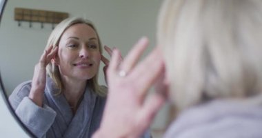 Banyodaki son sınıf beyaz kadın aynaya bakıyor, yüzüne dokunuyor. emeklilik yaşam tarzı, evde zaman geçirme..