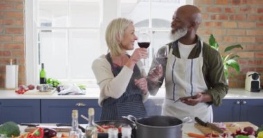 Karışık yarış çifti önlük giyip evde yemek pişirirken şarap içiyorlar. emekli çift yaşam tarzı konsepti