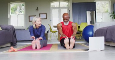 Karışık yarış son sınıf çifti evde dizüstü bilgisayara bakarak esneme egzersizi yapıyor. emeklilik ve aktif yaşlı yaşam tarzı konsepti