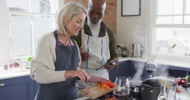 Evdeki mutfakta yemek pişirirken önlük giyip tablet kullanan son sınıf melez bir çift. emekli çift yaşam tarzı konsepti
