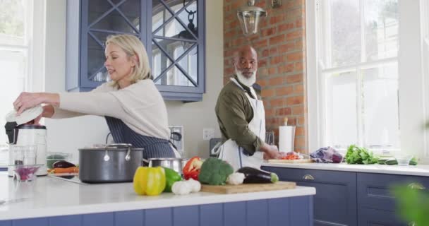 家庭で台所で一緒に調理エプロンを身に着けている混合レースのシニアカップル 定年退職したシニアカップルのライフスタイルのコンセプト — ストック動画