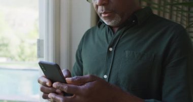 Afro-Amerikan son sınıf öğrencisi evdeki pencerenin yanında akıllı telefon kullanıyor. emeklilik son sınıf yaşam tarzı konsepti