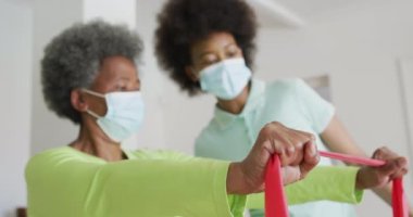 Yüz maskesi takan Afrika kökenli Amerikalı kadın fizyoterapist yaşlı kadın hasta egzersizlerine yardım ediyor. Üst düzey sağlık ve tıbbi fizyoterapi tedavileri, covid 19 salgını sırasında.