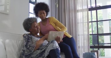 Afrikalı Amerikalı anne ve yetişkin kız kanepede oturup tablet kullanıyorlar. emeklilik hayatı teknolojiyle evde dinleniyor..