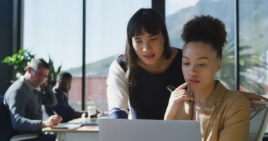 Modern bir ofiste çalışan iki melez iş kadınının ön planda dizüstü bilgisayar ekranına bakıp arka planda çalışan meslektaşlarıyla yavaş çekim tartışmaları.