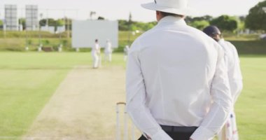 Arka tarafta bir kriket hakemi ve beyaz giyen genç bir melez erkek kriket oyuncusu, kriket maçında sahada bowling oynarken, arka planda diğer oyuncularla birlikte, yavaş çekimde.