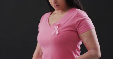 Göğsündeki pembe kurdeleyi siyah arka planda gösteren bir kadının orta kısmı. Göğüs kanseri farkındalığı kavramı