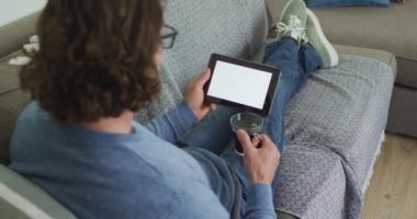 Kafkasyalı bir adam oturma odasında kanepede oturup tablet kullanıyor ve kahve içiyor. Yurtiçi yaşam tarzı, evdeki iletişim teknolojisini kullanarak..
