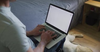 Beyaz adam evcil köpekle yatak odasında dizüstü bilgisayar kullanıyor. Yurtiçi yaşam tarzı, evdeki iletişim teknolojisini kullanarak..