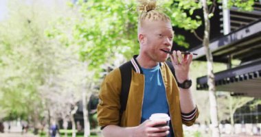 Parkta rastalı saçlı, kahve içen, akıllı telefondan konuşan mutlu albino Afrikalı Amerikalı adam. Dijital göçebe şehirde geziyor, dolaşıyor..