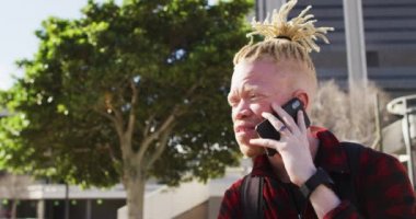 Parkta rastalı saçlı, akıllı telefondan konuşan mutlu albino Afrikalı Amerikalı adam. Dijital göçebe şehirde geziyor, dolaşıyor..