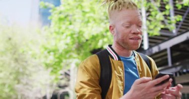 Parkta rastalı saçlı, akıllı telefondan konuşan mutlu albino Afrikalı Amerikalı adam. Dijital göçebe şehirde geziyor, dolaşıyor..