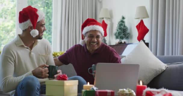 ノートパソコンのクリスマスビデオ通話中にコーヒーを飲みながら受け入れるサンタの帽子の幸せな父親と息子 クリスマスやお祭りや通信技術は — ストック動画
