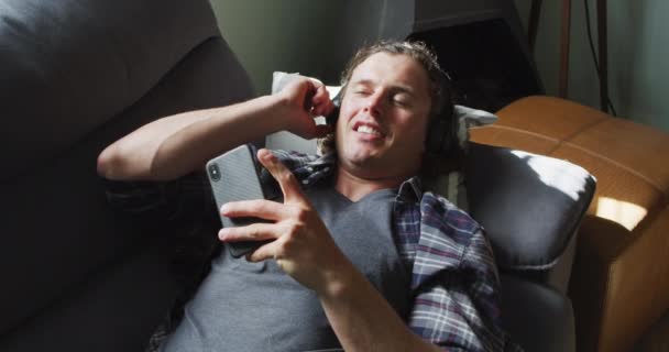 リラックスした白人男性がスマートフォンを使用し リビングルームでソファに寝そべってヘッドフォンを着用しています 家庭でのコミュニケーション技術を使って — ストック動画