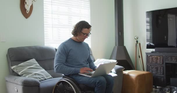 坐在轮椅上的快乐的高加索残疾人在客厅里使用笔记本电脑 有身体残疾的家庭生活方式 — 图库视频影像