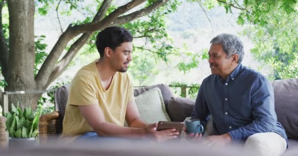 快乐的父亲和儿子坐在院子里的沙发上与平板电脑和咖啡杯聊天 家庭生活方式 饮品及通讯科技 — 图库视频影像