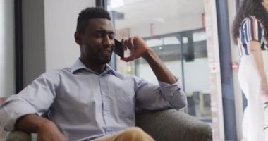 Gülümseyen Afro-Amerikan iş adamı koltuğunda oturmuş modern ofisteki akıllı telefondan konuşuyor. iş ve işyeri.