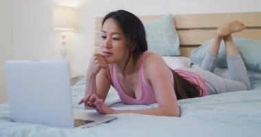 Mutlu Asyalı kadın sabahları dizüstü bilgisayar kullanarak yatakta yatıyor. Teknoloji ile evde yaşam tarzı ve rahatlama.