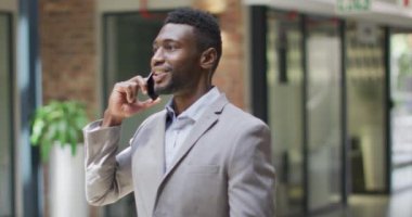 Gülümseyen Afrikalı Amerikalı iş adamı koridorda dikilip modern ofiste akıllı telefondan konuşuyor. iş ve işyeri.