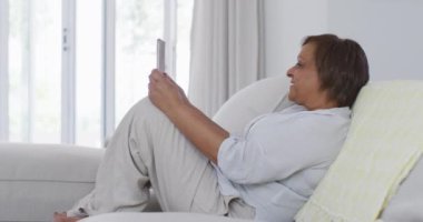 Mutlu Afro-Amerikan son sınıf öğrencisi kadın kanepede oturmuş tablet kullanarak video görüşmesi yapıyor, gülümsüyor. Emeklilik hayatı, evde teknolojiyle baş başa zaman geçirmek..