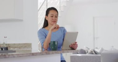 Masada oturan, kahve içen ve mutfakta tablet kullanan Asyalı kadın. Teknoloji ile evde yaşam tarzı ve rahatlama.