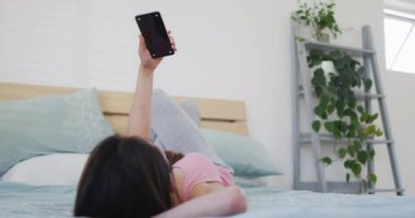 Asyalı bir kadın yatakta uzanıyor ve sabahları fotokopi makinesiyle akıllı telefon kullanıyor. Teknoloji ile evde yaşam tarzı ve rahatlama.