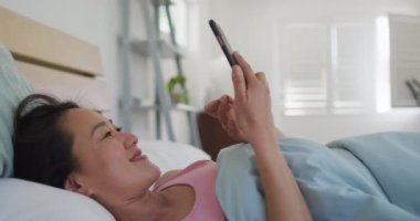 Mutlu Asyalı kadın sabahları akıllı telefon kullanarak yatakta yatıyor. Teknoloji ile evde yaşam tarzı ve rahatlama.