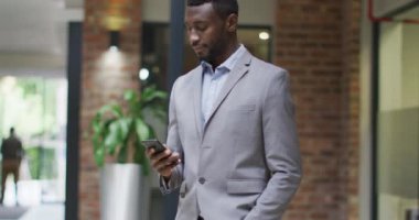 Gülümseyen Afrikalı Amerikalı iş adamı modern ofisteki akıllı telefonu kullanarak koridorda duruyor. iş ve işyeri.