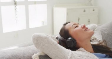Mutlu Asyalı kadın kanepede uzanıyor, kulaklıklarla evde dinleniyor. Teknoloji ile evde yaşam tarzı ve rahatlama.