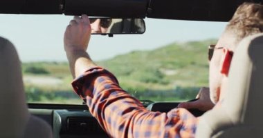 Güneş gözlüklü beyaz adam plajda güneşli bir günde arabanın içinde dikiz aynasını ayarlıyor. Yaz gezisi ve doğada tatil.