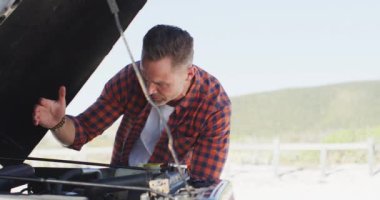 Stresli beyaz adam plajda güneşli bir günde kaputu açık arızalı arabaya bakıyor. Yaz gezisi ve doğada tatil.