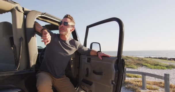 阳光明媚的沙滩上 戴着太阳镜的快乐的高加索人坐在开着车门的车里 暑期公路旅行及自然假期 — 图库视频影像