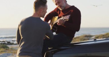 Arabasında oturup gitar çalan ve plajda konuşan mutlu beyaz erkek çift. Yaz gezisi ve doğada tatil.