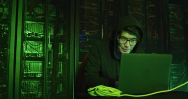 Bilgisayar sunucuları tarafından dizüstü bilgisayar kullanan kapüşonlu Asyalı erkek hacker. İnternet güvenliği, siber suç, bilgi teknolojisi, veri işleme ve bilgisayar sunucuları.