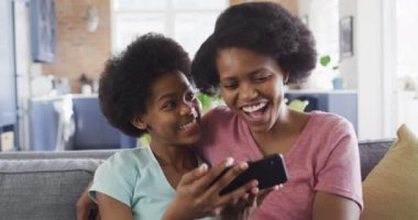 Mutlu Afro-Amerikan anne ve kızı akıllı telefon kullanarak kanepede oturuyor. evde aile hayatı ve kaliteli aile zamanı.