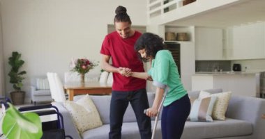 Rastalı gülümseyen melez bir adam oturma odasında kadın partneri tekerlekli sandalyeye oturtuyor. Fiziksel engelli refah ve ev hayatı.