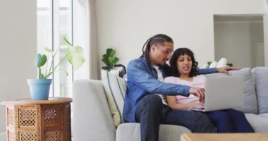 Çift ırklı mutlu çift kanepede dizüstü bilgisayar kullanarak oturuyor ve oturma odasında konuşuyor. Refah ve ev hayatı, evde birlikte dinlenmek..