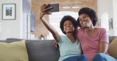Mutlu Afro-Amerikan anne ve kızı kanepeye oturmuş akıllı telefon kullanıyor ve selfie çekiyor. evde aile hayatı ve kaliteli aile zamanı.