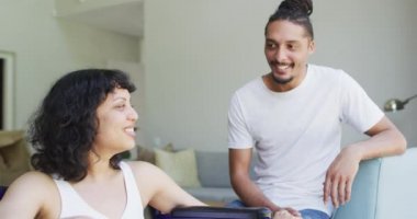 Tekerlekli sandalyedeki mutlu melez kadın ve oturma odasında birbirine bakıp gülümseyen erkek partner. Fiziksel engelli refah ve ev hayatı.