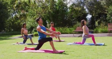 Çeşitli gruplar erkek eğitmenle yoga yapıyor, güneşli parkta minderlerin üzerinde diz çöküyor. fitness, sağlıklı hobiler ve açık havada aktif yaşam tarzı.