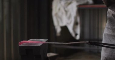 Beyaz erkek demircinin orta kısmı güvenlik gözlüğü takıyor, örse sıcak metal aletler takıyor. Demirci ocağında küçük bir işletme ve işçilik..
