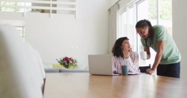 Tekerlekli sandalyedeki melez kadın ve erkek partner kahve içip oturma odasında dizüstü bilgisayar kullanıyorlar. Fiziksel engelli refah ve ev hayatı.
