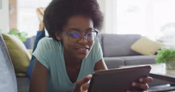 幸せなアフリカ系アメリカ人の10代の少女のソファの上に タブレットを使用して横たわっていた 家庭でのコミュニケーション技術を使って — ストック動画