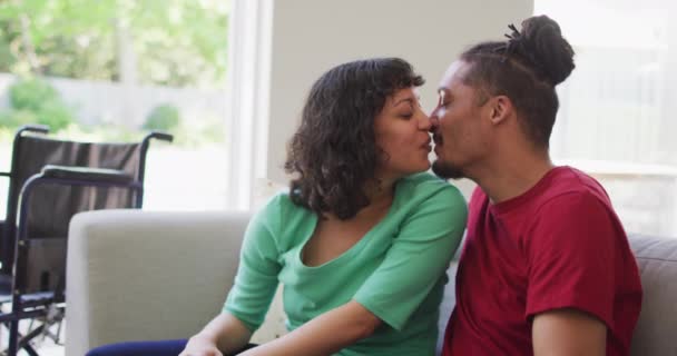幸せな出産のカップルソファのリビングルームでキスに座って 車椅子を背景に 身体障害者の健康と国内生活 — ストック動画