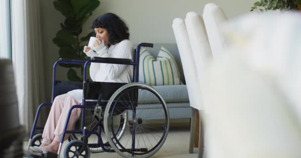 車椅子の女性がコーヒーを飲みながらリビングで窓から外を眺めていると 幸せな出産障害者の女性 身体障害者の健康と国内生活 — ストック動画