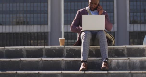 城市里的非裔美国人坐在楼梯上喝咖啡 用的是智能手机和笔记本电脑 城市里到处都是数字游牧民族 — 图库视频影像