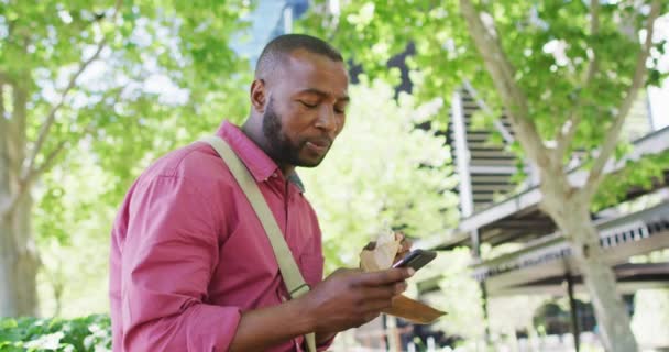 Afro Amerikan Adam Şehirde Yemek Yiyip Akıllı Telefon Kullanıyor Dijital — Stok video